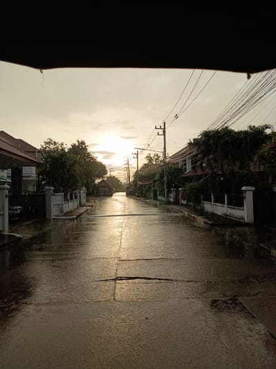 This media file depicts a street scene in Ban Patan, Thailand, during a rain shower. The time of day appears to be either early morning or late afternoon, with the sun low on the horizon, casting a warm, golden light that reflects off the wet asphalt. The rain is visible as streaks and droplets, creating puddles and a glistening sheen on the road.
The scene is lined with houses, some with red-tiled roofs, and lush green trees and vegetation. White fences and pillars, characteristic of the area, border the properties. Utility poles with a dense network of overhead wires are prominent along the street, a common sight in many parts of Thailand. The overall mood is somewhat melancholic due to the rain, yet the soft, diffused sunlight adds a touch of serenity. No people are visible in the image, suggesting the inhabitants are indoors, seeking shelter from the rain. The focus of the image is the street itself, glistening under the rain and the soft light, leading into the distance where more houses and trees can be seen. There is no visible text in the media file.