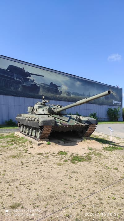 A green tank is parked outdoors on a sunny day in front of a building with a large mural depicting tanks. The tank is on a raised dirt platform, and it is facing to the right of the image. The mural behind the tank shows several tanks, one of which is firing its cannon. The building has a grey, corrugated metal exterior. The sky is a clear blue with a few small clouds. The ground in front of the tank is covered in sand and sparse vegetation. A small sign is visible to the right of the tank. The text on the mural in the upper right corner appears to be in Russian and reads "МИР ТАНКОВ". The timestamp in the bottom right corner reads "2026/7/15 10:32". The text in the bottom left corner reads "REDMI NOTE 9 PRO AI QUAD CAMERA". This appears to be an exhibit at a tank museum.