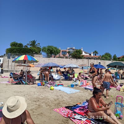 The image depicts a crowded beach scene in Trani, Italy, on a clear, sunny day. The beach is sandy, with numerous people relaxing on towels and beach chairs under umbrellas. Several individuals are seen in swimwear, some sitting, others standing or walking. Objects on the beach include beach bags, buckets, toys, and folded towels. In the background, there are stone walls, lush green trees, palm trees, and buildings with Mediterranean architectural styles, indicative of a coastal Italian town. The sky is a clear, bright blue. A timestamp in the lower right corner indicates "22.06.2026 10:54".