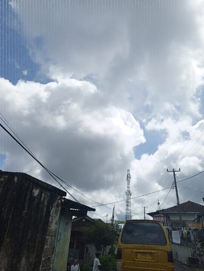 A cloudy sky looms over a street scene. A yellow van with license plate "F 1946 HA" sits on the road. Telephone poles and an industrial cell tower stand among the buildings. Clothes are hanging on a line outside one of the houses. Two people are standing in the lower-left corner. The buildings appear worn and weathered.