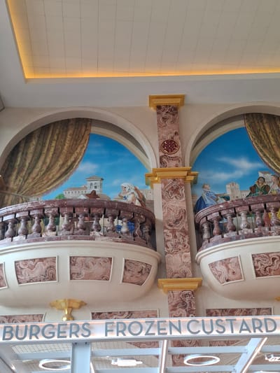 The image shows an interior view of a food establishment with a faux balcony mural. The mural depicts a scene with people on a balcony overlooking a town with a blue sky. The architecture in the mural and the decorative columns suggest a classical or historical theme. The foreground of the image features a sign that reads "BURGERS FROZEN CUSTARD" in illuminated lettering. This sign is suspended from a white structure, and below it, circular and linear lights are visible. The overall impression is of a themed dining area, possibly within a larger complex. The city of Trafford in the United Kingdom is mentioned as the location, suggesting this establishment is situated there.