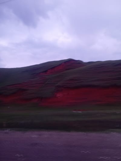 A hillside landscape under a cloudy sky. The hillside is covered in green vegetation with a prominent band of exposed red soil or rock running horizontally across its middle section. The sky is filled with dark, heavy clouds, tinged with purple hues, suggesting either twilight or an overcast day. In the foreground, there is a dark, textured surface that appears to be a road or path. The image does not depict any people, specific buildings, or identifiable landmarks that would confirm its location within Taldyk, Kyrgyzstan. The scene is natural and rural in appearance.