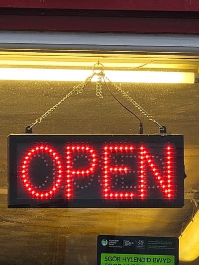 The image shows an "OPEN" sign hanging from chains in Holywell, United Kingdom. The sign is a black rectangular box with the word "OPEN" displayed vertically in bright red LED lights. To the left of the sign, a fluorescent light emits a bright yellow glow. To the right, a food hygiene rating sticker is partially visible. The background appears to be a textured surface, possibly a wall.