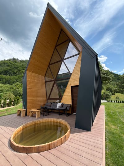 In Bistrica, Montenegro, a modern, angular wooden and dark-paneled house sits on a deck overlooking a grassy slope and a lush, tree-covered mountainside. In the foreground, a large, oval wooden hot tub filled with murky water is situated on the deck. The house features a striking triangular facade with a large, geometric window and a brown wooden door. On the deck, a dark-colored sofa with grey cushions and a white checkered throw is placed near two small, light-wood stools. The environment is characterized by bright daylight under a partly cloudy sky.