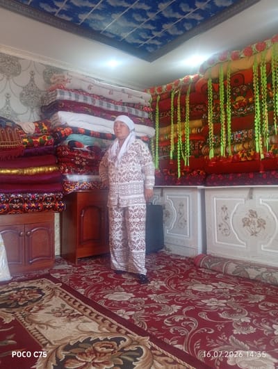 A woman stands in a room filled with stacked bedding and rugs. She is wearing a white headscarf and a matching patterned outfit. The room has a carpeted floor and walls covered in patterned wallpaper. A portion of the ceiling is decorated with a sky motif. In the foreground, the text "POCO C75" and a date "16.07.2026 14:35" are visible. The scene is likely a domestic interior in the city of Чоң-Гара, Kyrgyzstan.