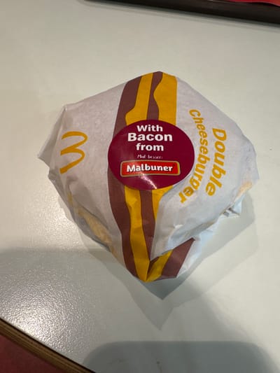 The image shows a McDonald's Double Cheeseburger with bacon, still in its wrapper. The wrapper is white with yellow and brown stripes, and yellow "Double Cheeseburger" text. A red circular sticker says "With Bacon from Malbuner." The McDonald's logo is visible at the bottom of the wrapper. The image appears to be taken on a table, likely inside or near a McDonald's restaurant. Spreitenbach, Switzerland is the presumed location, based on the presence of the German language "Malbuner" branding on the sticker.