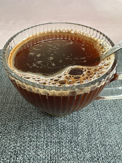 A close-up, overhead shot shows a clear glass mug filled with dark coffee. The coffee has a frothy head with many small bubbles. A silver spoon rests in the mug on the right side, with its handle extending out of frame. The mug sits on a textured gray surface, possibly a placemat or tablecloth. The background is a soft, muted pink. The image is well-lit and focuses on the coffee and mug. No specific location in Дольче Вита Кп, Russia, can be identified from the visual information provided.