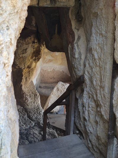The image depicts a view into a rock-cut cave or niche, possibly part of the Rock-hewn Churches of Ivanovo, Bulgaria, given the presence of deteriorated frescoes. The view is obstructed by a framework of wooden beams and support structures, seemingly added for preservation or stabilization. The walls and ceiling are roughly hewn from rock, exhibiting a textured, uneven surface. Faint traces of painted figures are visible on the interior walls, suggesting religious or symbolic iconography. The scene is dimly lit, with light entering from an external source, highlighting the textures of the rock and the details of the frescoes. The overall impression is one of age, fragility, and historical significance, with modern interventions aimed at preserving the site.