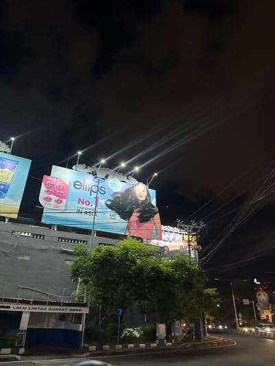 This is a nighttime outdoor photograph of a street scene in Denpasar Barat, Indonesia. The primary subjects are two large billboards advertising beauty products. The larger billboard features a woman with dark hair and a pink top, with the text "ellips No. 1 HAIR VITAMIN IN ASIA" prominently displayed. Below this, there's an image of pink hair vitamin capsules. The second, partially visible billboard below advertises "carasun" and shows a blue sunscreen bottle with "SPF 50+" and "15¢" visible.
The scene is illuminated by artificial lights from the billboards and streetlights, creating a dramatic contrast against the dark sky, which appears to be cloudy. The billboards are mounted on structures and appear to be alongside a road. In the background, there are trees, buildings with grey facades, and street signs. One sign reads "LALU LINTAS SUNSET ROAD". The overall atmosphere is commercial and urban, typical of a busy street at night. The photograph is taken at an angle, giving a dynamic perspective of the environment.