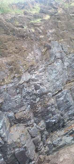 The image displays a rocky cliff face or outcrop situated in Ainamoj Village, Kenya. The rock formation consists primarily of dark grey to black sedimentary or layered igneous rock, characterized by visible stratification and fractures. Numerous sections of the rock surface appear wet or damp, reflecting light and exhibiting patches of white and light grey mineral deposits or efflorescence. Sparse green vegetation, consisting of grasses and small plants, is visible along the upper edge of the rock formation, indicating the ground level above. No individuals, animals, man-made objects, or discernible actions or interactions are present in the scene. The environment is natural and geological.