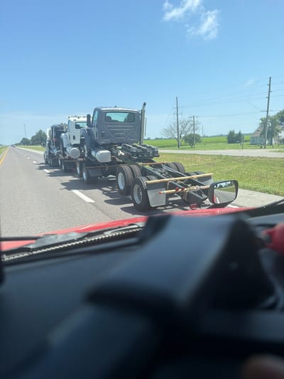 In Hayti, Missouri, United States of America, a convoy of trucks is captured on a sunny day on a two-lane highway. The trucks, all being transported, are arranged in a line, with the closest truck in the foreground appearing to be the last in the convoy. The trucks are being towed, with their rear axles and chassis visible. The environment appears to be rural, with green fields and scattered trees lining the sides of the highway. The sky is clear blue with a few wispy clouds. The photograph is taken from inside another vehicle, with the dashboard and windshield partially visible in the foreground.
