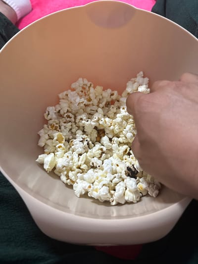 A hand reaches into a light pink bowl filled with popcorn. The popcorn is mostly white with some darker, burnt pieces scattered throughout. The bowl is sitting on a dark green textured surface, possibly a blanket. In the upper left corner of the image, a person's legs are visible, wearing white socks and a pink garment. The scene is likely indoors, given the soft lighting and close-up perspective. There is no identifiable information about the city of Fulda, Germany in this image.