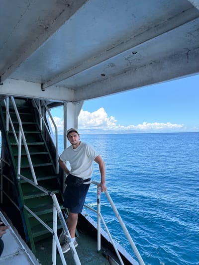 A man stands on an elevated walkway with white railings, likely part of a ferry or ship. He wears a light-colored T-shirt, dark shorts, a beige cap, and grey shoes. He holds a small, patterned object in his hand, possibly a cup. The background consists of the ocean and a blue sky with clouds. The structure the man is standing on has a green and white painted metal frame. The photo appears to be taken at a tilted angle. The city of Gunung Malang, Indonesia is not visibly discernible in the image.