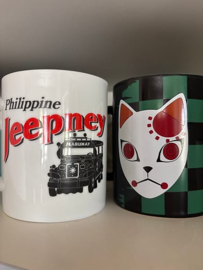 The image displays two mugs stacked vertically. The top mug is black with a green checkered pattern and features a white cat face design with red accents for eyes, nose, and ears. The bottom mug is white with black text and graphics. The text "Philippine" is printed vertically on the left side of the mug in a stylized font, followed by "Jeepney" in larger, red lettering. To the right of the text is a black silhouette of a jeepney, a common mode of transportation in the Philippines, with "MABUHAY" printed vertically on its side. The scene is likely indoors, given the lighting and the plain background. The image captures a close-up view of the mugs, with a shallow depth of field. There are no people visible in the image. The overall impression is a display of merchandise.
