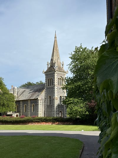 This is a vertical, medium-wide shot of a stone church with a tall spire, seen through the leaves of an ivy-covered structure. The church is made of grey stone with white decorative trim around the arched windows. The spire is pointed and made of a lighter stone, with decorative elements along its length. A sloped, grey tiled roof is visible on a lower section of the church. Lush green trees and bushes surround the building, and a well-maintained lawn with a paved path runs alongside it. The sky is clear and blue, suggesting a bright, sunny day. The image appears to be taken in the daytime, possibly late morning or early afternoon, given the lighting. There is no visible text in the image. The scene evokes a sense of tranquility and historical significance, typical of a church setting in a well-kept garden. The ivy in the foreground adds a natural, slightly overgrown element to the composition.