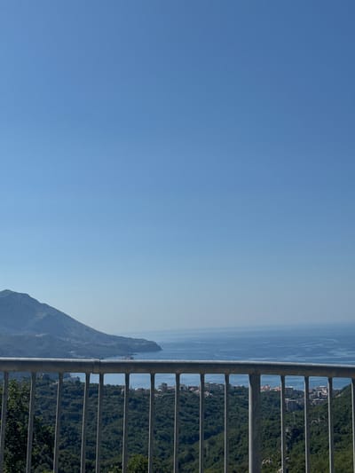 The image captures a serene vista from a high vantage point in Markovići, Montenegro. In the foreground, a sturdy metal railing with vertical bars separates the viewer from the landscape below. Beyond the railing, lush green trees cover the slopes, dotted with buildings that suggest a settlement nestled within the foliage. The middle ground reveals a coastline with the vast expanse of the Adriatic Sea stretching towards the horizon, where it meets the clear blue sky. A prominent mountainous peninsula extends into the sea on the left, showcasing its rugged terrain and the subtle hints of human habitation along its shores. The overall scene is characterized by a tranquil atmosphere, bathed in the bright light of a sunny day, with a hazy effect in the distance as the sea meets the sky.