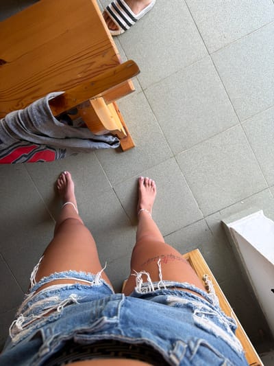 An overhead shot shows a person's legs and feet from the knees down. The person is wearing ripped denim shorts and white anklets. They are barefoot, with light pink nail polish on their toes. The person is seated on a wooden bench, with a grey t-shirt draped over it. The bench is placed on a grey tiled floor. In the upper right corner of the image, a foot wearing a black and white striped sandal is visible. A white structure is partially visible in the lower right corner. The image does not provide any specific details to indicate the location in the city of Chavei, Bulgaria.