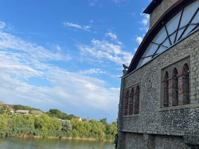 A stone building with arched windows and a large triangular window is situated next to a body of water. A small statue of a dog stands on the roof of the building. The water is bordered by lush green vegetation, and in the background, there are houses and trees. The sky is blue with scattered white clouds. The image is captured from a slightly elevated perspective, looking across the water towards the building and the opposite shore.