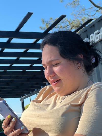 A medium shot shows a young woman with dark hair pulled back in a messy bun and fair skin looking down at her phone. She is wearing a light yellow t-shirt and a silver necklace. She has painted nails in white with yellow accents, and rings on her fingers. She is outdoors, with a dark wooden pergola structure in the background and a bright blue sky above. Trees are visible in the distance, along with part of a building. The scene appears to be in a public outdoor space in the city of Ochamchire, Abkhazia.