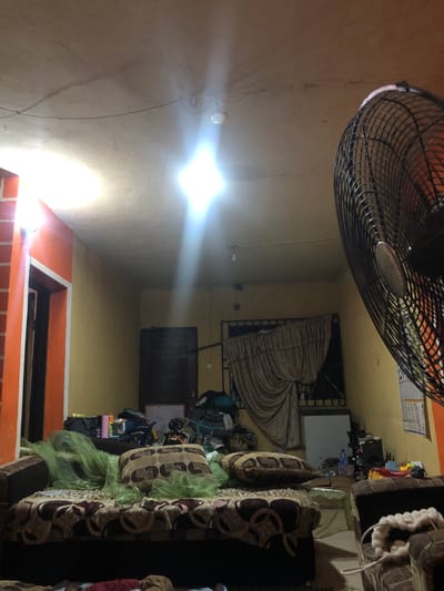 A room with a ceiling fan dominating the upper portion. A bright ceiling light casts a strong glare. A sofa runs along the right side, its patterned cushions visible. Curtains cover a window. Items are scattered around a whiteboard near the center. A door is visible in the background. Below, a red-and-orange-tiled structure is apparent.