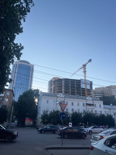 The image shows a street scene in Novopodkletnoye, Russia, bathed in bright daylight, suggesting it was taken in the morning or afternoon under clear skies. The orientation of the image is vertical.
The primary subjects are several buildings under construction and completed structures, along with parked cars and trees. A prominent element is a large construction crane positioned next to a multi-story building in the early stages of construction, with its skeletal frame exposed. Adjacent to this, a modern, glass-fronted skyscraper stands tall. Other older buildings with traditional windows are also visible.
Parked along the street are several cars, including a dark-colored sedan and a white hatchback. A few traffic signs are present: a triangular warning sign and a blue circular sign with a white arrow indicating a turn. The street appears to be paved.
Trees with dark green foliage line parts of the street, providing a contrast to the urban architecture. The sky is a clear, bright blue, with a few small birds visible. Power lines crisscross the scene, adding to the urban feel.
One sign visible on a building appears to have Russian text, with "ПЛЮС" prominent on a red background. Another sign further down the street also shows Cyrillic script, possibly indicating a business or service. The overall impression is of an active urban environment, with ongoing development and daily life.