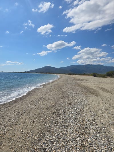 A wide, pebble-strewn beach stretches along the coast of Astros, Greece, under a bright blue sky dotted with white, fluffy clouds. The sea, a deep blue, gently laps at the shore, creating a thin line of white foam. In the distance, rolling green mountains form a scenic backdrop, their peaks softened by the haze. The beach itself is a mix of sand and countless small, smooth pebbles in various shades of grey, white, and brown. A few scattered green bushes and low-lying vegetation can be seen along the edge of the beach, hinting at the natural landscape of the region. The scene is serene and peaceful, evoking a sense of a quiet summer day by the sea.
