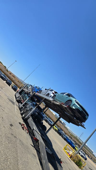 This is a low-angle, full-shot photograph taken on a bright, sunny day in Pedrola, Spain, focusing on a car carrier truck loaded with vehicles. The truck is positioned on an asphalt surface, likely a parking lot or transport hub, with a clear blue sky dominating the upper portion of the frame. The truck is angled, with its front end pointing upwards and to the left, showcasing multiple tiers of cars being transported.

On the top tier of the carrier, closest to the viewer and most prominently featured, is a light teal-colored car with a sleek, modern design, characterized by its distinctive headlights and wheels. Behind it, other cars are visible, including a white SUV and a blue sedan. The lower decks of the truck are also loaded with dark-colored cars, some partially obscured by the upper levels.

In the background, a row of parked cars is lined up neatly, displaying various colors including blue, yellow, and white. A yellow metal barrier structure is visible in the middle ground, and further in the distance, there are buildings and power lines, suggesting an industrial or commercial area. The environment is dry and arid, with some sparse vegetation visible on a hillside in the far left background. A tall light pole stands on the left side of the frame, and a metal pole with a white top is on the right. The overall scene depicts a logistics operation involving the transportation of new vehicles.