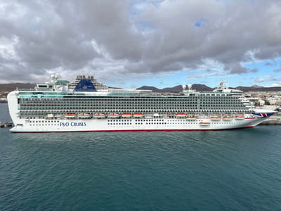 A large white P&O Cruises ship, identified by "P&O CRUISES" text on its hull and a blue funnel with a gold sunburst logo, is moored in the harbor of Puerto del Rosario, Spain. The vessel features a red waterline stripe, multiple decks lined with windows and balconies, and numerous orange lifeboats along its side. The ship's bow displays a Union Jack flag design. The surrounding water is deep blue-green with slight surface ripples. In the background, a low-rise city with light-colored buildings stretches along the shore, backed by arid, undulating hills under an overcast sky featuring scattered grey clouds. No individuals or specific actions are discernible in the scene.