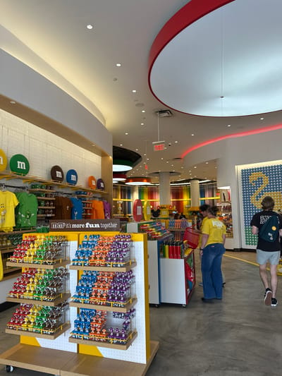 This is an interior shot of the M&M's store located in Lake Buena Vista, Florida, United States. The store's design includes curved white ceilings with red accents and recessed lighting. A column with colorful M&M displays and dispensers is centrally located in the store. People are browsing the merchandise, with a couple in the upper right corner and another individual in a yellow shirt seen with a red shopping basket. Display racks filled with M&M's branded merchandise, including t-shirts and small figures, are located in the foreground. A large sign reads "M&M’s it’s more fun together," emphasizing the brand's message. Overall, the store has a bright, colorful, and playful atmosphere, fitting with the M&M's brand identity.