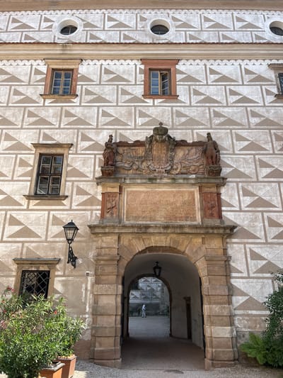 This is a medium-angle, eye-level shot of the Sgraffito courtyard entrance at Náchod Castle in Náchod, Czechia. The facade is adorned with a repeating geometric sgraffito pattern in shades of white and beige. An arched stone entryway dominates the lower center of the frame, leading to a courtyard visible in the background. Above the arch, a large, ornate stone crest with a crown and figures is mounted on the wall. Two stone statues stand on either side of the crest. A large inscription is carved into a stone panel below the crest. To the left of the arch, a tall, dark lantern with a decorative bracket is attached to the wall, positioned above a window with a black iron grate. A lush green plant in a planter sits in the lower left corner of the image. The overall impression is one of historical architecture and intricate design.
