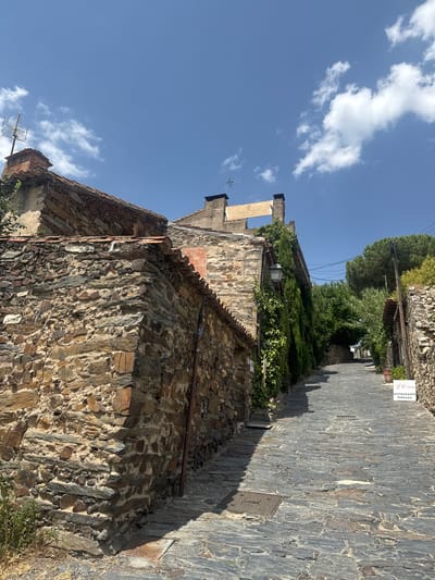The image shows a view looking up a cobblestone street in the city of Patones, Spain. The street is narrow and lined with traditional stone buildings. On the left side of the street, a large stone wall is prominent, with parts of a building constructed with reddish-brown tiles and brickwork visible above it. A chimney with an antenna is also seen on the rooftop. Green ivy and other plants climb the walls of the buildings. On the right side of the street, a sign for "La Cabaña RESTAURANTE TERRAZA" is visible, along with terracotta pots and dense greenery. The street itself is paved with irregular grey cobblestones, and a manhole cover is present in the foreground. The sky above is blue with scattered white clouds, indicating a sunny day. The overall impression is of a charming, historic village street scene.