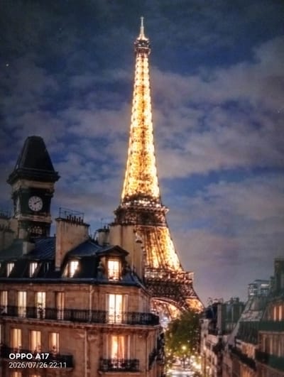 The image is a night-time view of the Eiffel Tower in Paris, France. The tower is illuminated and stands tall against a cloudy blue sky. In the foreground, there are Parisian-style buildings with illuminated windows and balconies. A clock tower is visible to the left of the Eiffel Tower. The streets below are also lit, suggesting an urban environment. The scene is a classic postcard view of Paris. The location of Құмөзек, Kazakhstan is not depicted in this image.