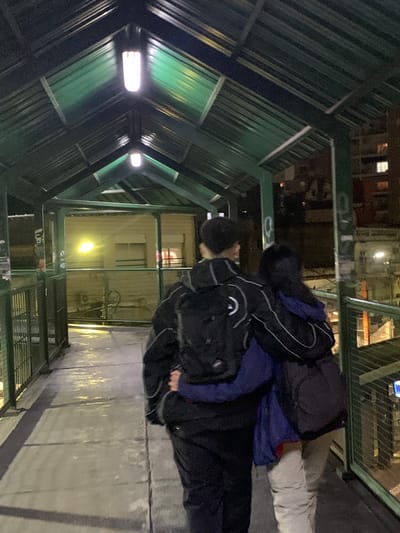 Two individuals are observed on a walkway, potentially a pedestrian overpass or elevated platform. The primary subject, closer to the viewer, is wearing dark clothing and carrying a dark backpack. The second individual, partially obscured, is wearing a dark jacket with blue accents and light-colored pants. The walkway is constructed of concrete or a similar material. The environment suggests an urban location in Ramos Mejía, Argentina, indicated by the building architecture and the overall lighting. The scene takes place at night, with artificial lighting sources illuminating the area. The structure above the walkway features a corrugated metal roof.
