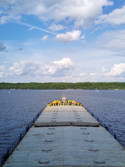The image shows the deck of a barge traveling on a body of water, presumably a river or lake, with a forested shoreline in the distance. The sky is blue with scattered cumulus clouds.

The deck of the barge is a gray, flat surface with rows of circular hatches. Blue railings line the sides of the deck. Towards the far end of the barge, a yellow structure with various equipment is visible.

The water is calm with slight ripples, reflecting the blue of the sky. The distant shoreline is densely forested.

The location is Novodevich'e, Russia. This information is based on common knowledge of barge transportation on Russian waterways.