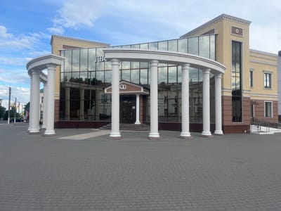 The image shows the front facade of a two-story building with a classical architectural style, located in the city of Bobrov, Russia. The building has a modern glass facade in the center, flanked by traditional beige-colored sections with brown trim. A semicircular colonnade with white pillars and a matching archway frames the main entrance. The entrance itself is a small portico with double doors and a sign above it. The ground in front of the building is paved with gray bricks. The sky is partly cloudy. There are no people visible in the image. The reflection in the glass reveals details of buildings across the street. A ramp is on the right side of the building.