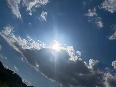 The image shows a bright, sunny day with a blue sky filled with scattered clouds. The sun is positioned high in the sky, partially obscured by a large, dark cloud, casting visible sun rays. On the left side of the frame, a dark, silhouetted hillside covered in vegetation is visible against the bright sky. The image does not contain any people, specific objects, or actions indicative of the city of Prijepolje Municipality, Serbia. The scene is primarily focused on the natural landscape and sky.
