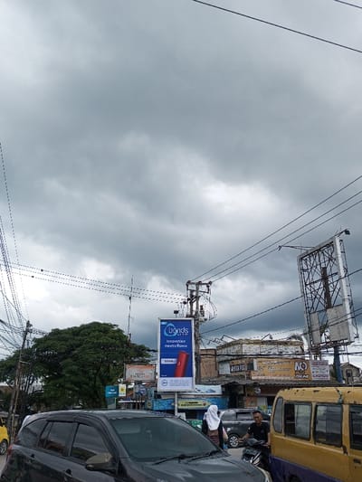 The image presents an urban street scene in Cibadak, Indonesia, viewed under a heavily overcast sky. In the foreground, a dark-colored multi-purpose vehicle (MPV) occupies the left half, angled slightly right. A woman wearing a white hijab and dark clothing is walking from left to right behind this vehicle. To the right, a man in a black shirt is seated on a black scooter, facing the camera, positioned next to a yellow minibus with a distinct purple lower section.

Above the street level, a dense network of overhead power lines and communication cables spans the frame, connected to a central utility pole equipped with multiple transformers and junction boxes. To the right of the utility pole, a vertical blue billboard advertises "Bonds by S.M." with the text "Innovation meets flavor" and an image of a red cylindrical object, alongside a "21+" age restriction. Various commercial buildings with storefronts and signage, some bearing Indonesian text like "MANDIRI" and "TOKO," line the street in the mid-ground. A large, leafy green tree is visible on the far left, behind the utility infrastructure. On the far right, a large, empty billboard frame stands prominently against the cloudy sky.