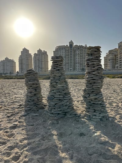 In Al Jazeerah Heritage Village, United Arab Emirates, three towers constructed from stacked, flat stones stand on a sandy beach. The sun, low in the sky and casting long shadows, illuminates the scene. In the background, a row of modern, multi-story buildings with distinctive architectural features are visible, contrasting with the natural elements in the foreground. The beach is comprised of sand mixed with shells and small pebbles. The sky is clear and a pale blue.