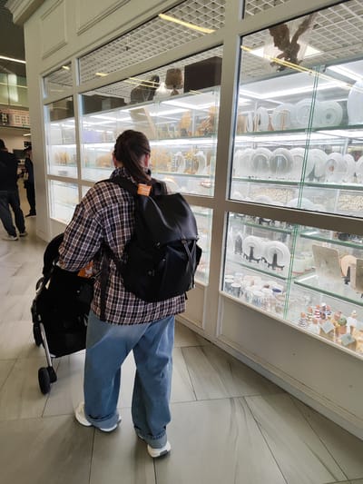 The image captures an interior view of what appears to be a duty-free shop at Khrabrovo Airport in Kaliningrad Oblast, Russia. A woman, seen from behind, is walking with a stroller. She is wearing a plaid shirt and blue jeans, with a black backpack on. The shop displays various items on illuminated shelves behind glass, including what seem to be plates, cups, and decorative figurines. The flooring is tiled with a marble-like pattern. To the left, two men are visible in the background, one wearing a dark jacket and the other in a dark shirt. A sign with Russian text is also visible on the left wall.