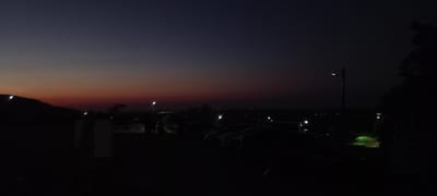The image depicts a low-light scene, likely during twilight or dusk, in the vicinity of Gara Oreshets, Bulgaria. The sky transitions from a deep blue at the top to hues of orange and red near the horizon, indicating the setting sun. In the foreground, several vehicles are parked in what appears to be a parking area. The vehicles are dark silhouettes against the dimly lit environment. A street lamp is visible to the left of the frame, casting a faint light. To the right, a tree's dark foliage is prominent. In the distance, faint lights suggest the presence of a settlement or urban area. The overall atmosphere is quiet and subdued, typical of a rural or semi-rural location at this time of day. No people are clearly discernible in the image.