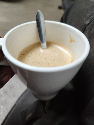 A close-up, overhead shot shows a white ceramic mug filled with frothy coffee. A metal spoon is partially submerged in the coffee. The mug is held by a person's hand, visible on the left side of the frame. The background is out of focus, showing dark fabric, possibly clothing or upholstery. The lighting appears to be ambient and diffused. The scene does not provide any discernible information about its location in the city of Volevčice, Czechia.