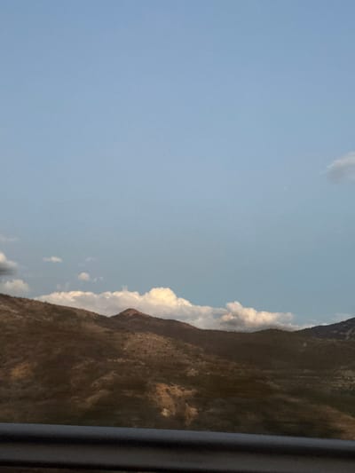 A bright blue sky dominates the view, punctuated by a few scattered clouds. Along the right side, a rugged, brown mountain range stretches upwards, its peak partially obscured by a line of puffy white clouds. The photo is taken from inside a vehicle, as indicated by the presence of a blurred black vertical line, potentially a car frame or window pillar.