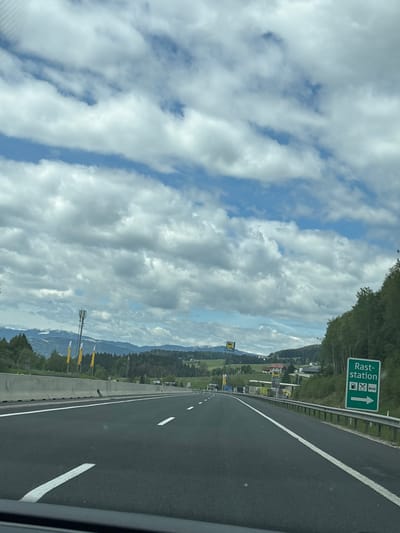 The image depicts a highway in Unterauerling, Austria, under a partly cloudy sky. A green road sign indicates a rest stop ("Rast-Station") with symbols for gas station, restaurant, and restroom, and an arrow pointing forward. The highway is black asphalt with white lane markings. Trees and hills flank the road. In the background, mountains are visible. A cell tower is located on the left. The photo was likely taken from inside a vehicle.