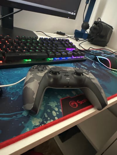 A close-up, slightly angled, eye-level shot shows a grey camouflage patterned gaming controller resting on a blue and green patterned mousepad. The controller is oriented horizontally and is the primary subject. Behind the controller, a black mechanical keyboard with RGB lighting is visible, with some keys illuminated in red, green, and purple. To the right of the keyboard, a black gaming mouse with a neon pink and blue light strip is partially visible. In the background, a computer monitor is positioned, and a black headset is resting on a blue stand. The scene is indoors, likely a desk setup for gaming, and there is no indication of the city of Madara, Bulgaria.