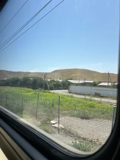 The image depicts a rural scene viewed through a curved window, likely from a vehicle. The dominant subjects are a dry, barren hillside on the left and a lush, green field on the right. The hillside is sculpted and devoid of significant vegetation, suggesting an arid climate. In contrast, the field is vibrantly green and appears to be a cultivated crop, possibly corn or similar, with rows clearly visible.

Between the hillside and the field, several man-made structures are present. There are white or light-colored buildings with corrugated roofs, suggesting agricultural outbuildings or simple dwellings. Power lines are visible running across the sky and also along the edge of the field, supported by utility poles. A chain-link fence runs along the right side of the image, separating the field from a gravel path or road.

The sky is a clear, bright blue, indicating a sunny day. The time of day appears to be midday or early afternoon, given the bright sunlight and lack of long shadows. The overall atmosphere is one of rural simplicity and agricultural activity, set against a backdrop of natural, rugged terrain.

There is no visible text in the image. The scene is characteristic of a location in Almachi, Uzbekistan, with its dry hills and agricultural fields. The image captures a moment of travel, with the curved window framing the passing landscape.