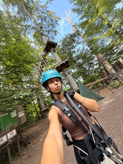 A young woman, wearing a blue helmet, brown top, and black shorts, with a harness, is in a treetop adventure park. She is taking a selfie with a pout. The scene is outdoors, with trees and wooden structures of the adventure park visible in the background. The sun is shining, casting shadows, suggesting it is daytime. There are signs visible in the background with German text and symbols indicating rules or information for the park.