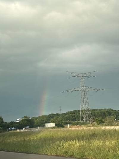 The image shows a rural scene near Dambach-la-Ville, France. A gray sky dominates the frame, with a faint rainbow visible. Overhead power lines and pylons traverse the landscape. A road runs along the right side of the image, bordered by grass and trees. Signage is visible beside the road, bearing town names and distances.