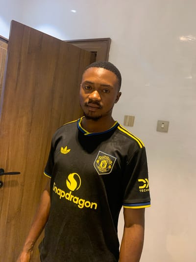 An adult male subject is depicted indoors, located in the city of Dong, Nigeria. The individual has dark skin, short dark hair, and a neatly trimmed beard, and is looking directly at the camera with a neutral expression. He is wearing a black, short-sleeved Manchester United jersey. The jersey features a yellow Adidas logo on the upper right chest, the Manchester United crest on the upper left chest, a prominent yellow "Snapdragon" logo across the mid-chest, and a yellow "DX Technology" logo on the right sleeve. The jersey also displays yellow stripes over the shoulders and blue and yellow trim on the sleeve cuffs.
The background to the subject's left consists of a light brown wooden door or paneling, with a black handle visible on the left side. To the subject's right, a plain white wall is visible. Mounted on this wall are two electrical wall plates: one off-white multi-gang plate with three vertical segments, and a separate, off-white single switch plate with a toggle switch, positioned slightly below and to the right of the first. The ceiling above is white.