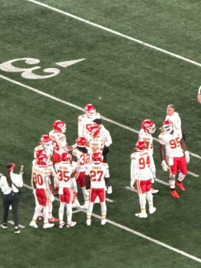 The image depicts an American football field, specifically the 50-yard line area, at MetLife Stadium in East Rutherford, United States. The artificial green turf features white yard lines and the numeral "50." A group of approximately ten Kansas City Chiefs football players is visible, wearing white jerseys with red numbers (e.g., 27 CORNEY, 32, 35 WATSON, 80, 94 HERRING, 95), red pants, and white helmets with red accents. They are standing in a loose formation, some facing each other. Two male non-player staff members are among the players; one wears a white long-sleeved shirt and black pants, and another wears a white shirt with a red bandana. A female staff member, wearing a white long-sleeved shirt, black pants, and a red baseball cap, stands on the left edge of the frame, holding a green and orange bottle. No active play is occurring; the individuals appear to be in a huddle or discussion during a break.