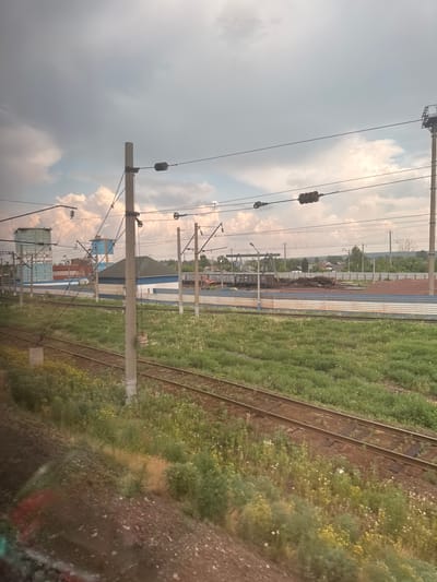 This is a photograph taken from a moving vehicle, likely a train, looking out at an industrial area and railroad tracks. The sky is overcast with dramatic clouds, suggesting a possibly stormy or humid day.

In the foreground, two sets of railroad tracks run diagonally across the frame, heading from the lower left towards the upper right. The tracks are surrounded by overgrown green vegetation, with patches of wildflowers visible amongst the grass. The ground on the far left is rough, dark earth with some scattered rocks and what appear to be remnants of discarded material.

Further in the mid-ground, there is a large, flat, blue and silver corrugated metal structure running parallel to the tracks. Beyond this, various industrial buildings are visible. These include at least two tall, rectangular structures with a blue and white color scheme, one appearing to be a water tower or silo. There are also smaller buildings with green and blue roofs, and piles of industrial materials, possibly construction debris or scrap metal. In the distance, a fence line is visible, with trees and more buildings beyond it, suggesting a rural or semi-rural setting bordering the industrial zone. A single orange piece of machinery, possibly an excavator, is visible near the fence line.

Several tall poles with electrical wires and insulators are spaced along the landscape, indicating an electrified railway system or power lines servicing the industrial area. One prominent pole stands in the center right of the frame, with multiple wires stretching across the image. The lighting suggests it is daytime, but the overcast conditions create a somewhat diffused light.

The image is characteristic of a scene found in an industrial or transportation hub. Given the context of Mariinsk, Russia, this scene could represent an area near the city's railway station or a manufacturing district. The presence of industrial infrastructure, railway lines, and varied vegetation is consistent with the broader landscape of such locations.